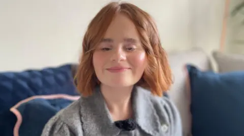 Evie Roberts has short wavy red hair, and is wearing a grey woollen top. She's sitting on a sofa with dark blue cushions.