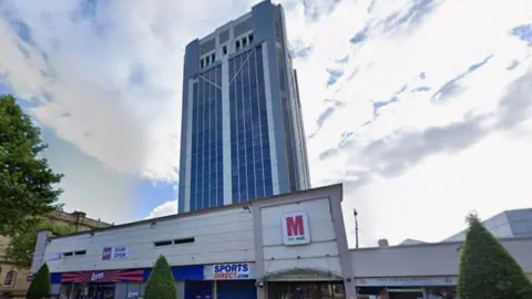 Street view image of the 16-storey white and grey clad tower block, rising about longer two storey shops and an entrance to the Mall shopping centre