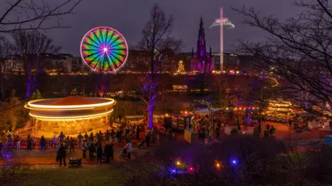 Kevin Carr Edinburgh's Christmas market, with stalls, a carousel lit up in yellow and a big wheel in the distance with green and red lights.
