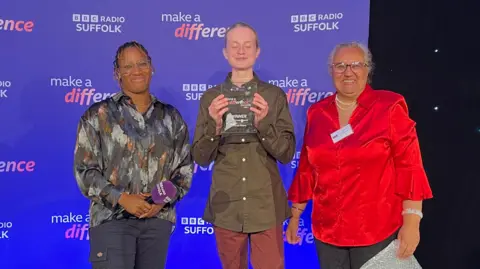 Seth Spain/BBC Angelle Joseph, James de Mierre and Gulshan Kayembe stand on a stage. James is holding a glass trophy. The backdrop says BBC Radio Suffolk Make a Difference