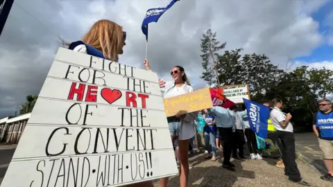 A picket line outside a school. There are several people standing holding banners and placards.
