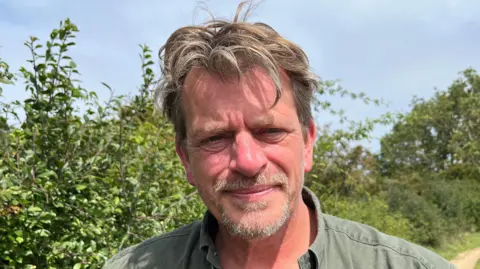 A man stands outside on a sunny day in front of a hedgerow. He has light brown hair some of which hangs over his forehead. He wears a green shirt. 
