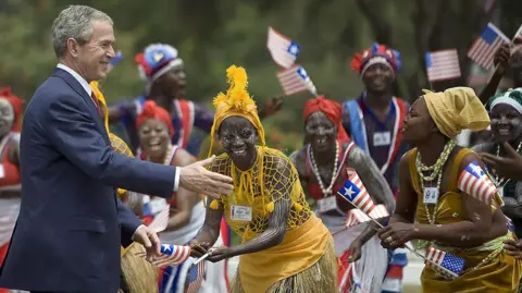 Former US President George W. Bush wearing a black suit reaching out to dancers who are wearing traditional attire and holding Liberian flags 