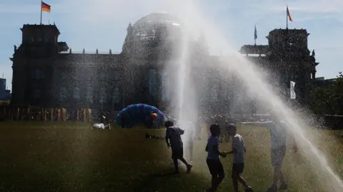 People pass by a watering system in front of the Reichstag on a hot summer day on June 24, 2026 
