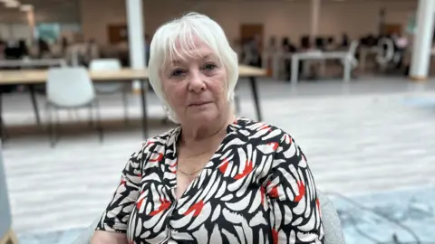 Kate Bradbrook/ BBC Danielle Stone in a white, red and navy patterned dress sits in a grey chair looking at the camera with lots of tables and chairs in the background