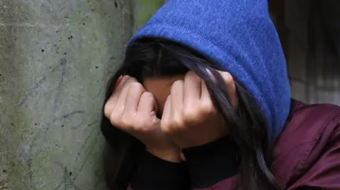 PA Media A teenage girl, wearing a blue hoodie, hides her head behind her hands which are clenched in fists.