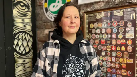 Simon Thake/BBC A woman in a black and white checked jacket stands in front of a brick wall with framed beer mates