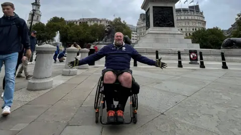 Chris Davis Chris with his arms outstretched whilst sat in a wheelchair. He is in Trafalgar Square. He is smiling at the camera.