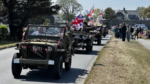 A cavalcade of vehices driving along a street. The vehicles have Union Jack flags attached to them. People are standing on the side of the road as they watch the vehicles drive past. It is a sunny day.