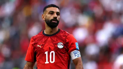 Mohamed Salah, visible from waist up, stands and looks tired during a game against Iran at the 2026 World Cup. He wears a red Egypt jersey with black trim, a white number 10 in the centre of the chest, a white sponsors logo and the crest of the Egyptian FA. Salah also has a white captain's armband around his upper left arm which has blue text which reads "football united" on it. The background is a heavily blurred crowd