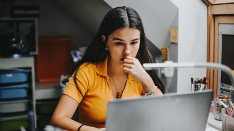 Getty Images A young woman in an orange top looks at her laptop while chewing her finger