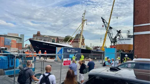BBC A trawler being harboured next to a wall surrounded by people in orange high vis and white helmets. A metal fence can be seen with people stood behind it looking.