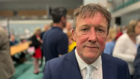 Neil Stevens wearing a white shirt, a pale blue tie and a navy blue jacket at teh election count held at Riverside Leisure Centre in Exeter