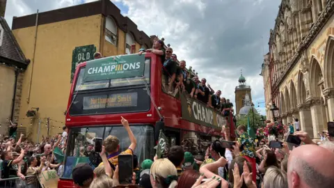 A red open-top bus with a green 'champions' banner' on it, surrounded by thousands of fans.