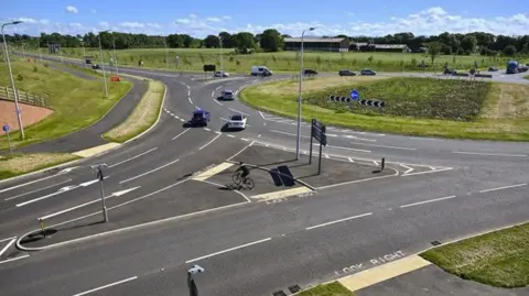 Cumberland Council An aerial image of the Newby West roundabout which has been enlarged and changed as part of the Carlisle Southern Link Road works. Cars are driving along the stricture, which is surrounded by green grass and trees. A cyclist is also using one of the crossings.