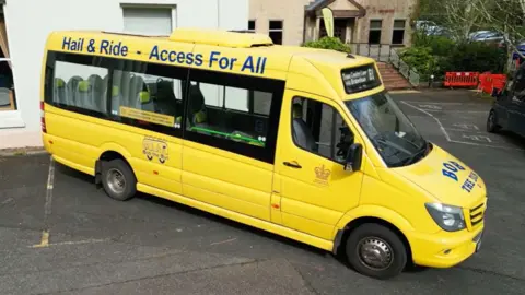 South Hams District Council A minibus-style vehicle in bright yellow is parked on a paved area outside a light‑coloured building. It is branded “Bob the Bus”, and also features a slogan “Hail & Ride – Access For All” printed in blue along the side.