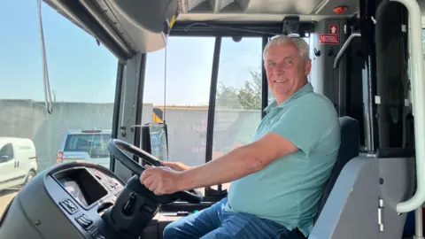 Henry Godfrey-Evans/BBC A man with a light green shirt in the driver's seat of a bus with his hands on the wheel