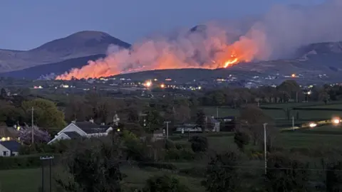 Más allá de las casas de campo, los setos, los árboles y las colinas onduladas, la cima de una montaña negra arde lentamente, con columnas de humo y fuego que se elevan hacia el cielo.