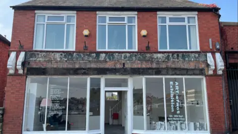 Megan Hayward The front of a red brick building. It has large windows with white windowpanes. Writing on the windows reads "ART-efact exhibition"