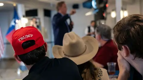 Attendees of a Bill Paxton rally watch him speak. Two of them wear hats; one of which is a signature red Trump cap