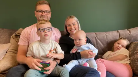 The whole family are on their sofa and looking at the camera. Dad James, mum Emily and their children Max, Rory and Holly.