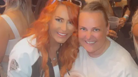 Kirsty Langley Kirsty Langley, 36, who has long red hair and is wearing a white and black jacket, poses for a picture with Shelley James, who is a similar age, has short blonde hair and is wearing a white t-shirt. The two are in the crowd at an arena and other concert goers are visible in the background. 