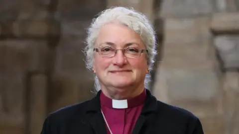 The Right Reverend Sarah Clark is in the middle of the image. She has grey hair in a ponytail and is wearing clerical clothing.