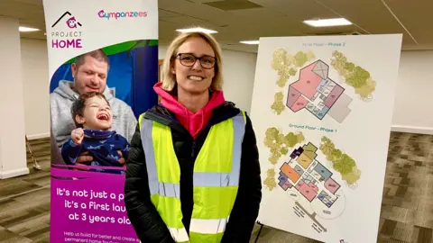 A person with short blonde hair and dark rimmed glasses smiling at the camera wearing a hi-vis jacket, standing in front of plans for the new building