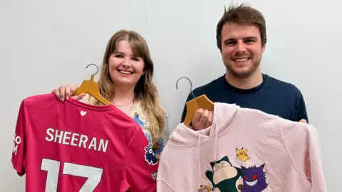 East Anglia's Children's Hospices Two people standing in front of a white wall. On the left is Bethany Watchman who is a woman with long brunette hair. She is looking directly at the camera and is smiling while holding up a bright pink football shirt which says in white writing SHEERAN 17 on the back. On the right is Scott Campbell who has short brown hair and is looking directly at the camera and smiling. He is wearing a blue T-shirt and is holding up a pale pink hoodie to the camera. Both items have been donated by Ed Sheeran and will be sold by the EACH charity.