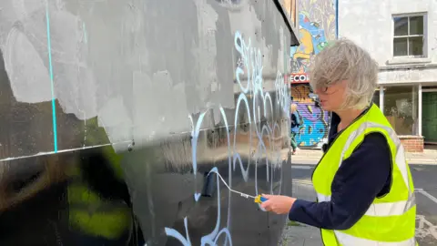 George Carden/BBC Emma wearing a high vis painting a black wall which has blue graffiti on it
