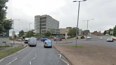 Cars and vans coming up to a roundabout with buildings in the background