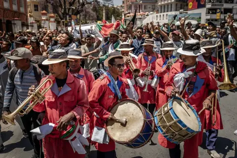 LUIS TATO / AFP / GETTY IMAGES Protesters dance and smile behind a marching bad who wear matching red clothes.