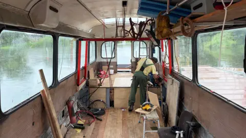 A wide shot of the interior of the bus boat. Paddy is seen in his green dungarees and yellow T-shirt with his back to the camera in the middle distance bending over and fixing something. The interior of the bus has a wooden floor and there are various pieces of wood and tools sitting around.