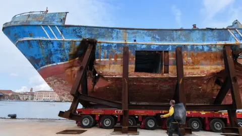 Getty Images The fishing vessel "Barca Nostra" (Our Ship) that sank on April 18, 2015 trapping hundreds of migrants in its hull, is being installed in Venice's former shipyards as part of the centerpiece of a new art project by Swiss-Icelandic artist Christoph Buechel, prior to the the 58th International Art Exhibition of the Venice Biennale, on May 7, 2019 in Venice. - The 58th International Art Exhibition will open to the public from May 11 to November 24, 2019.