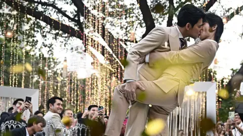 This photo taken on January 10, 2025 shows Thai actors Apiwat "Porsch" Apiwatsayree (L) and Sappanyoo "Arm" Panatkool in beige tuxedos sharing a kiss at their unofficial wedding ceremony in Bangkok. They are surrounded by family and friends taking photos of them under a tree decorated with twinkling lights.   