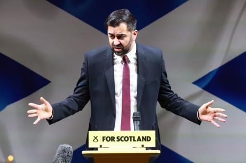 First minister Humza Yousaf standing at a podium making a speech with a Saltire flag backdrop