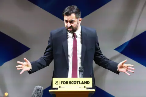 PA Media First minister Humza Yousaf standing at a podium making a speech with a Saltire flag backdrop
