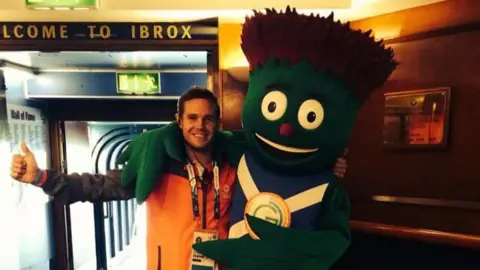 Sam Spittle Sam Spittle, a man with a brightly coloured top and a lanyard hanging around his neck, along with Clyde - an anthropomorphic thistle wearing a top with a Saltire pattern. The duo are standing next to a tunnel leading to a football pitch, with a Welcome to Ibrox sign hanging over it.