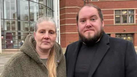 Parents of Wynter Andrew, Sarah and Gary. Sarah is wearing a khaki, fluffy hooded jacket with her hair tied in a pony. Gary is wearing a black wool coat.
