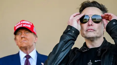 Donald Trump, wearing a Maga hat, stands behind Elon Musk, who is adjusting his sunglasses