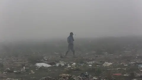 Getty Images A person walks through fog over rubbish-strewn ground while holding a hand over his mouth
