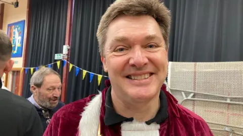 Jeremy Fish smiles at the camera inside a community hall. He has short dark hair and is wearing a red and white Santa Claus top. Behind him other volunteers at a wrapathon event can be seen talking.