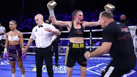 In a boxing ring in full kit, Emma Dolan raises her hands in victory next to her coach, as she is announced to have earned a split decision victory in Birmingham, over a disconsolate opponent, Shannon Ryan, pictured left.