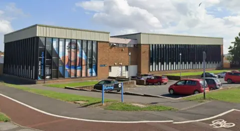 Google Streetview of the existing Woodhouse Close Leisure Complex. It is large and square with narrow long windows, one wall of which is covered with a picture of a woman posing in a swimming pool. There us a small car park next to it.
