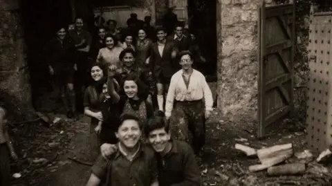 Black and white picture of children walking towards the camera. They walk outside the castle, with brickwork visible and sticks and wood on the floor.