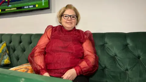 Maria smiles at the camera as she sits on a green sofa which is right next to a white wall. There is part of a green table in front of her and two pillows to her left. She has a red, silk top on and is wearing glasses. Maria has short blonde hair.