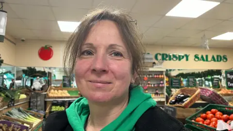 Trudie looking into the camera, she's wearing a green hoodie with a black fleece over the top. Behind her you can see rows of fresh fruit and vegetables, and a sign on the wall reads 'crispy salads'.