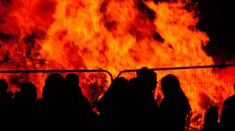 The picture shows a number of people silhouetted in front of a large bonfire. fencing is in front of the fire