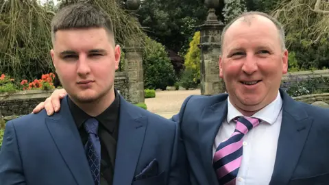 Joe Dix Foundation Joe Dix wore a black shirt, navy embossed tie and matching pocket chief, with a navy jacket. His father has his arm around him and is also wearing a navy suit with white shirt and a navy and pink striped tie. They are standing in front of an ornate wall, and gate opening, with well manicured gardens behind them too.