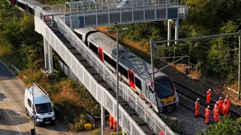 Jonathan Brady/PA A small group of people in orange hi-vis clothing and white hard hats stand on train tracks in front of a train that is slightly leaning to one side near a railway crossing bridge